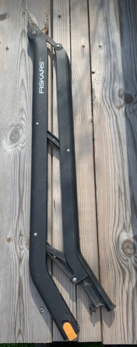 Fiskars sekatör i svart med enhands- och tvåhandsfattning ligger på trägolv.