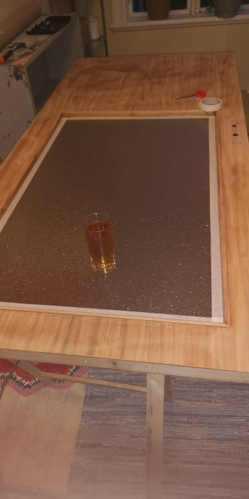 Ommålad träinnerdörr liggande på ett bord med ett glas på dörrytan och målarredskap i bakgrunden.