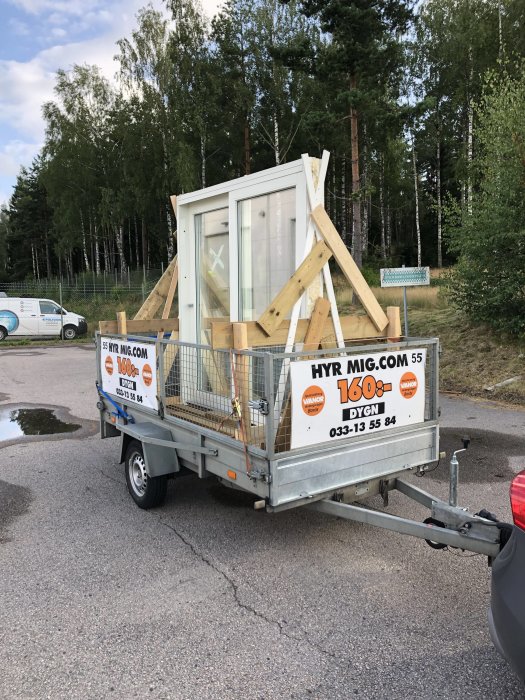 Släpvagn lastad med fönster och trästöttor säkrade med nät, parkerad på en asfalterad väg.