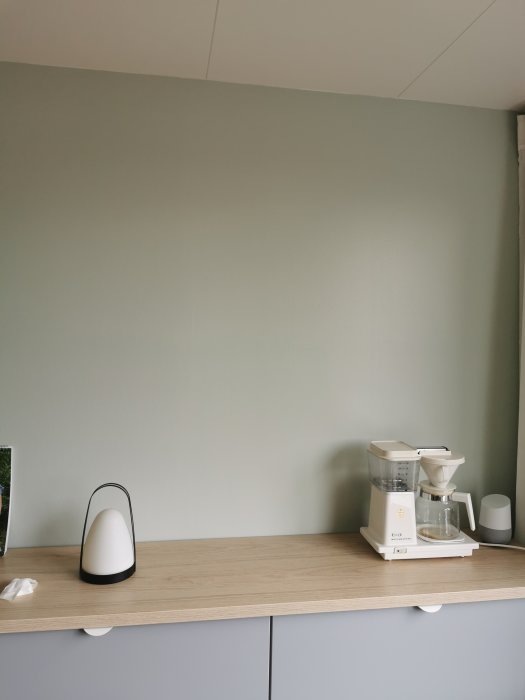 Vägg målad med Jotun-färgen "soft mint", kaffebryggare och lampa på köksbänk.