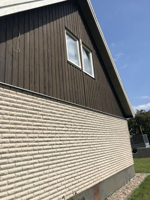 Del av villa med nedre fasad i rundhugget mexitegel och övre delen i mörk träpanel med två fönster.