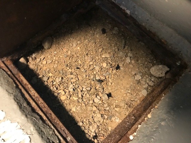 Tätt packat grus och sten i en rensbrunn i en källare, med synliga tecken på fukt och separering från betongplattan.