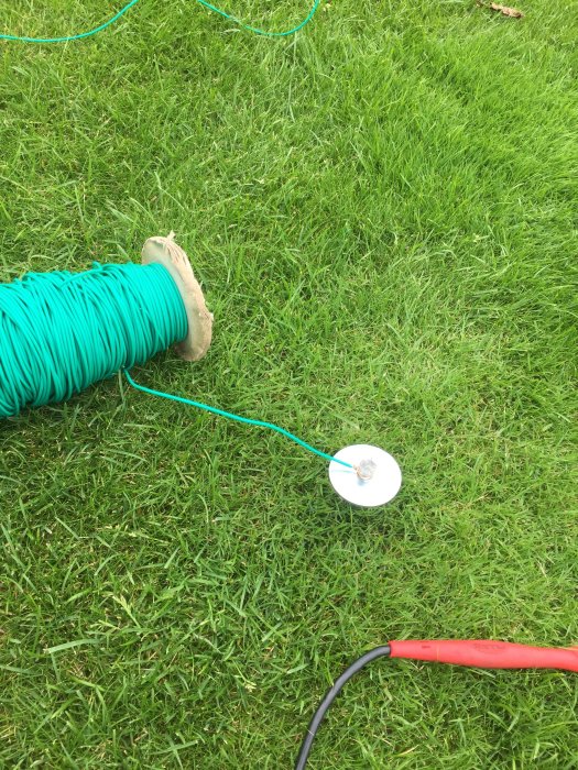 En rulle med grönt kabel på gräsmatta, jordspett och en del av en röd mätkabel.