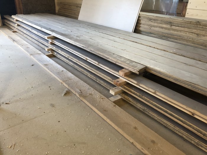 Stapel av återanvända golvplankor upplagda på träklossar i ett renoveringsprojekt.