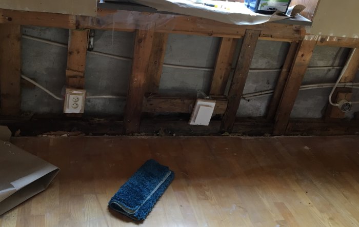 Skador på en vägg under ett fönster med synlig syll och isolering samt en blå trasa på trägolv.