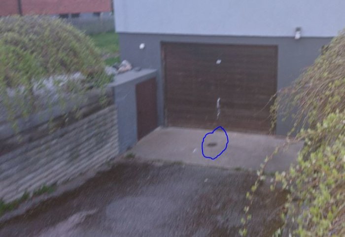 Uteplats med gråa betongväggar och en brun garageport, med en markerad blå cirkel där en brunn tidigare varit.