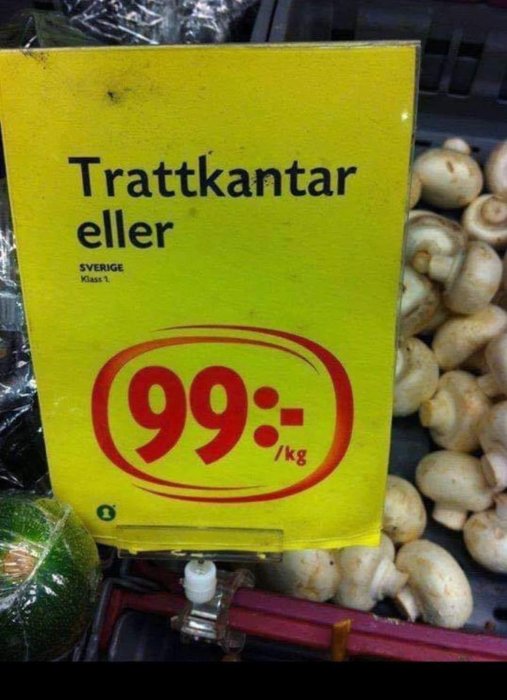 Prisskylt för trattkantareller till 99 kronor per kilo med champinjoner i bakgrunden.