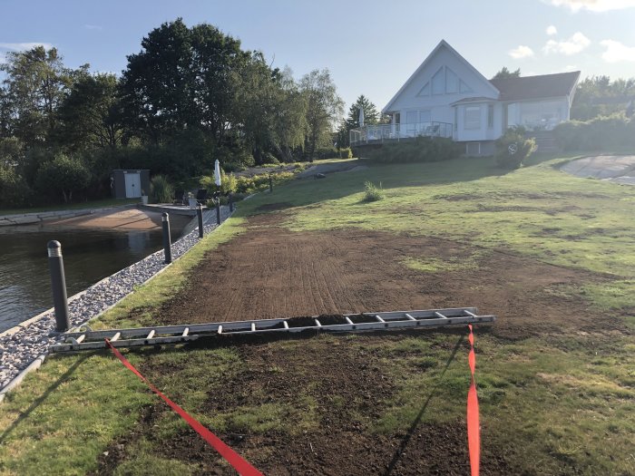 Vit villa med nyjordad sluttande trädgård och en brygga vid vatten.