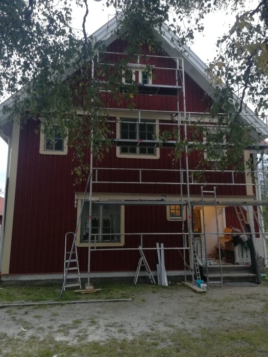 Röd träfasad på hus med uppsatt byggställning och stege, läkten monterad och klart för målning.