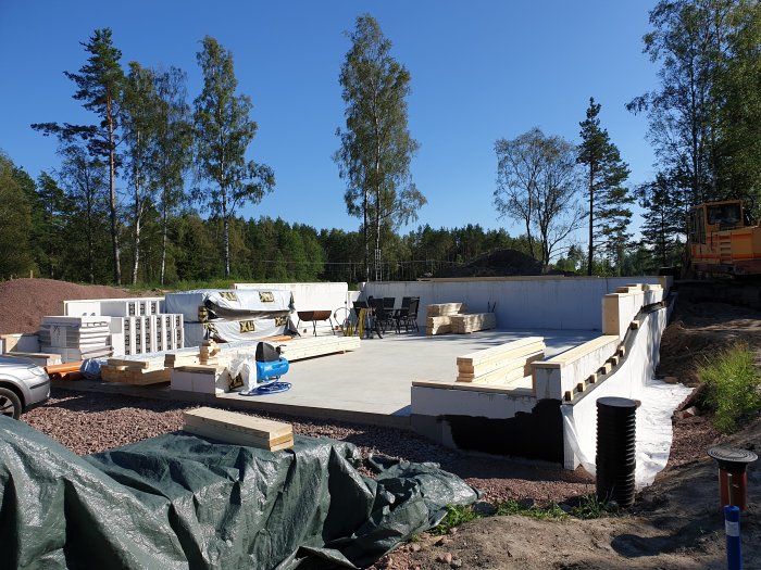 Byggplats med grunden till ett hus, trämaterial, isolering och en grävmaskin i bakgrunden under en klar himmel.