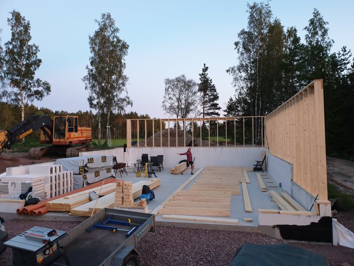Byggarbetsplats med uppförda väggar, trämaterial på marken och en grävmaskin i bakgrunden.