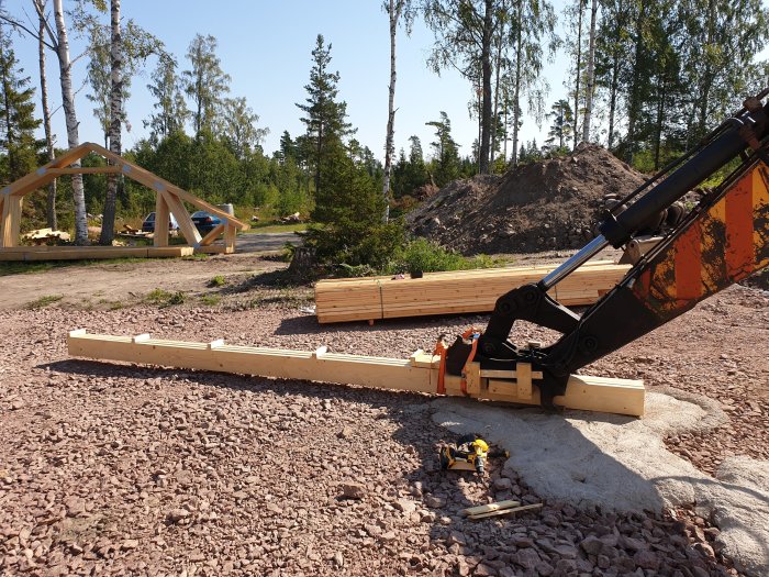 En grävmaskins arm som håller en improviserad lyftbalk framställd av plankor på en byggarbetsplats, med takstolar i bakgrunden.