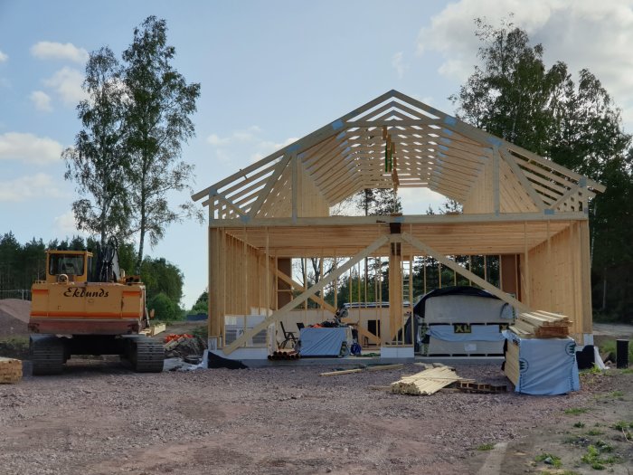 Uppförande av trästomme till byggnad med grävmaskin i förgrunden och delvis uppsatta takstolar.