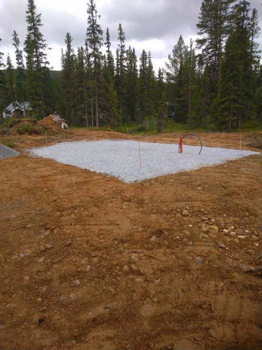 Grundläggning för förråd med nyligen påförd makadam, markerade hörn och skog i bakgrunden.