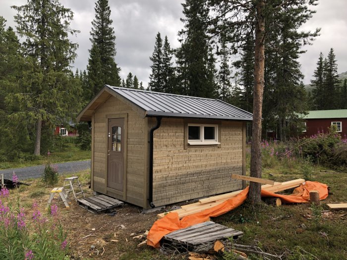 Förråd med nytt plåttak och råspontsklädsel i en skogsmiljö, byggmaterial syns i förgrunden.