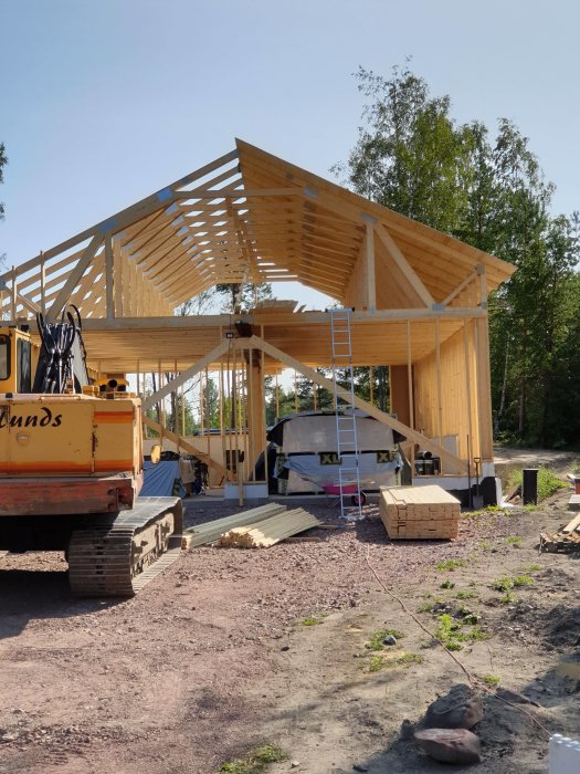 Delvis konstruerat trägarage med synliga takstolar och en stege, omgivet av byggmaterial och en grävskopa.