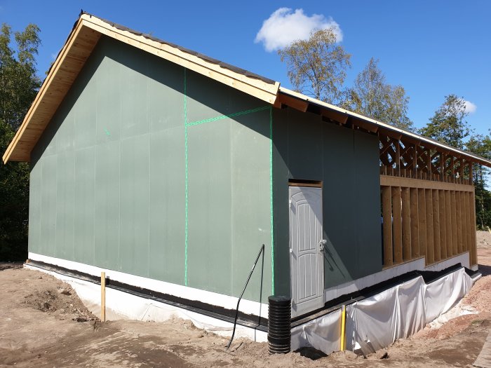 Ett delvis byggt garage med grön ytterbeklädnad, en vit dörr, och en loftgång under konstruktion, omgivet av sandig mark.
