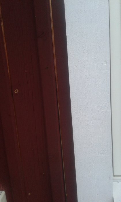 Spricka vid spik i mörkröd ytterväggsfoder intill vit karm, potentiellt orsakat av fuktinträngning.