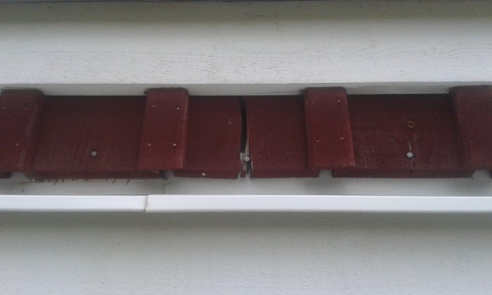 Närbild på rödfärgade ytterväggspaneler med en spricka nära en spik och en vit list som bakgrund.