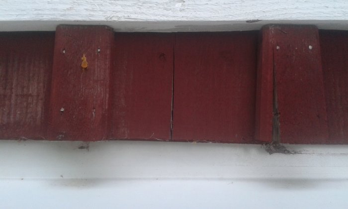 Sprickor runt en spik i en rödmålad ytterväggspanel nära vit fodrande list.