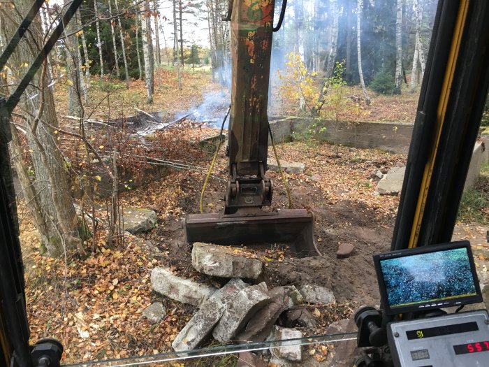 Vy från grävmaskinens hytt som gräver bort gammal husgrund omgivet av skog och höstlöv.