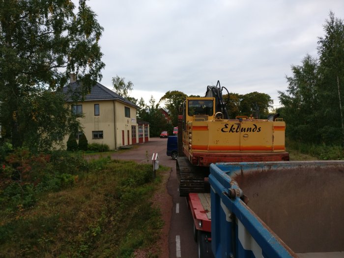 Gul grävmaskin på lastbil framför igenväxt tomt och bostadshus, möjlig framtida infart.