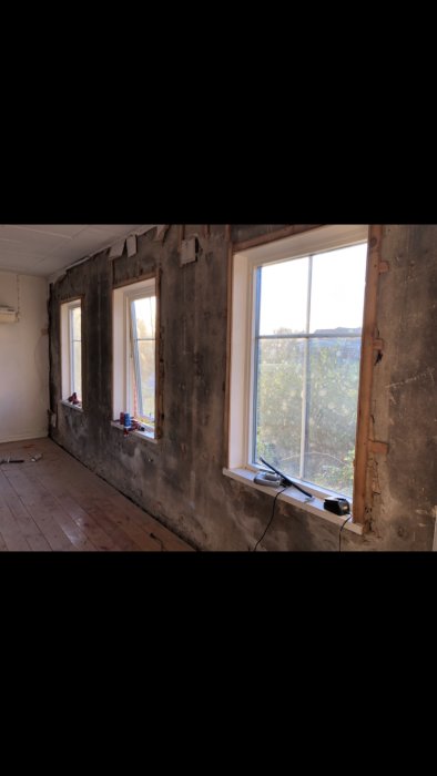 Interiör vy av ett renoveringsprojekt, visar ett rum med väggar där puts har skalats av och två fönster syns, redo för isolering.
