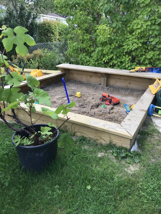 Nybyggd sandlåda i trä fylld med sand, leksakstraktor och spadar i en trädgård.