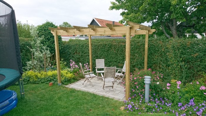 Nybyggd träpergola i trädgården med fyra stolar på stenplattor, omgiven av grönska och blomster.