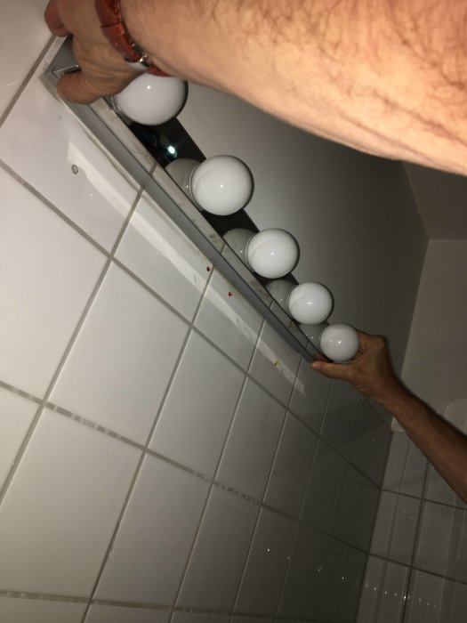 Person som håller en badrumslampa med vita kulor mot kakelvägg, inga synliga skruvar.