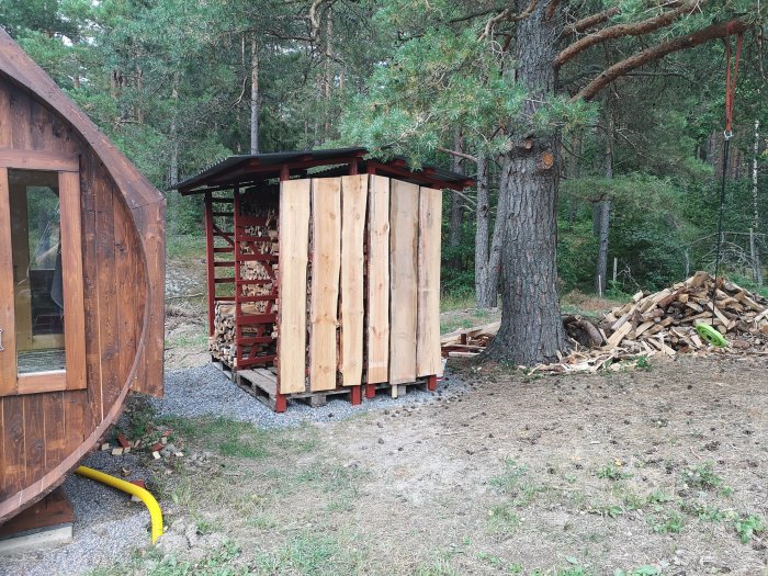 Ny flyttad vedstapel intill bastu, med återanvänt virke, intill en hög med kluvet ved.