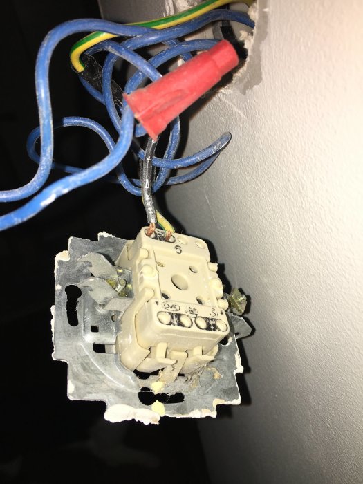 Strömbrytare med öppnat lock och synliga ledningar, en märkt med svart tejp, nära en vägg.