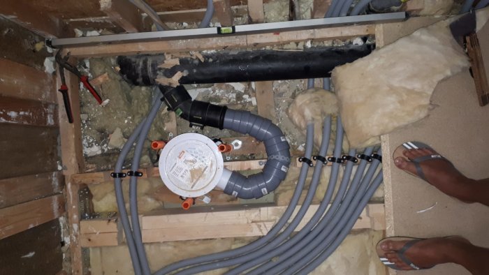 Vy över golvbjälklag med synliga rörinstallationer och isolering, tagen från ovan där en persons fötter ses.