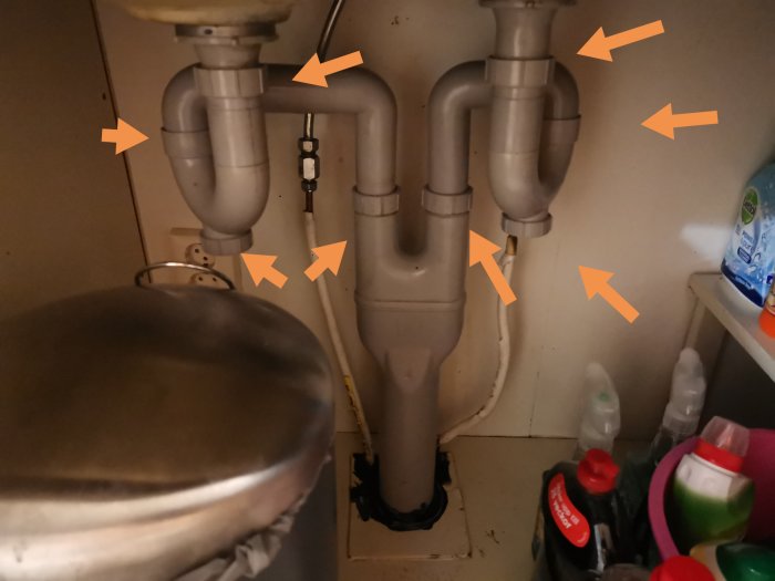 Under diskbänk med rörinstallationer och orangea pilar som pekar ut delar, omgiven av städprodukter.