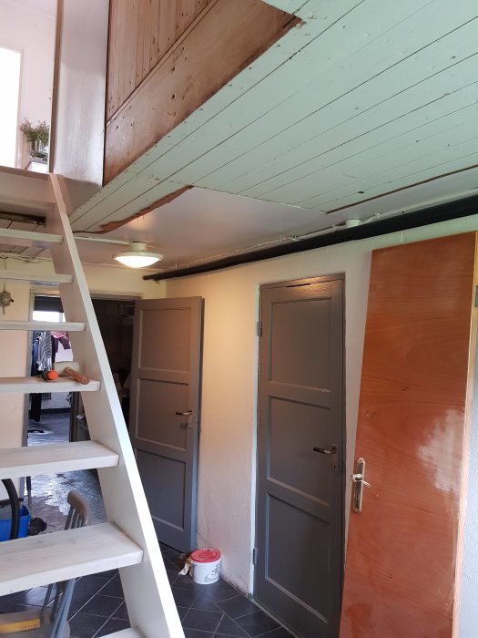 Bild på ett källarutrymme med en träbalk längs taket vid trapphål, grå dörrar och vit trappa.