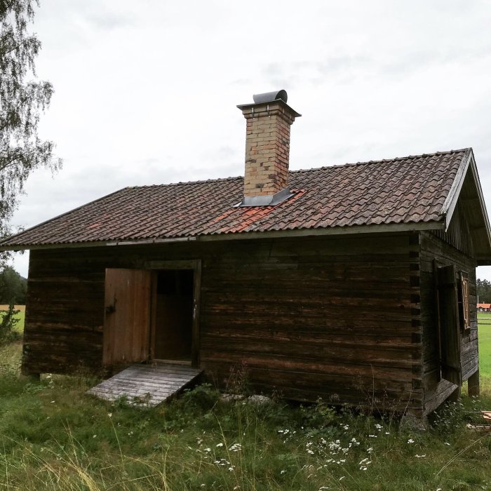 Gammal trästuga med slitet tegeltak och skorsten, öde och i behov av renovering omgiven av grönska.