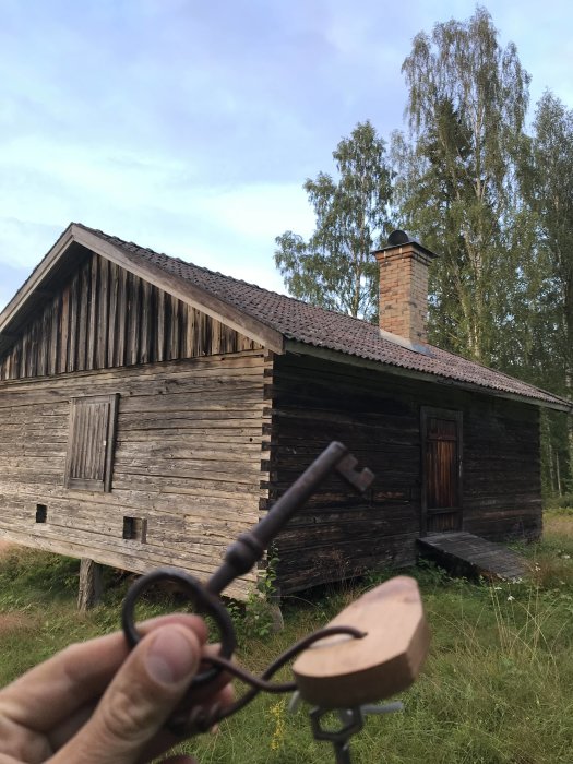 Hand håller en gammal nyckel framför en äldre stuga i behov av renovering.