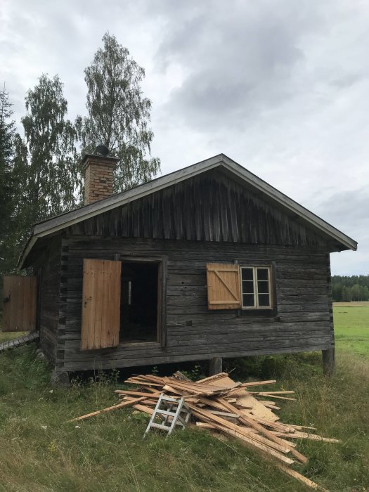 Äldre fäbodstuga i trä med öppen dörr och fönsterluckor, renoveringsvirke framför, omgiven av natur.