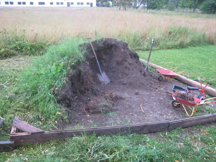 Grävd grop i en trädgård med två skottkärror, spadar och en hög med jord intill ett fält av långt gräs.