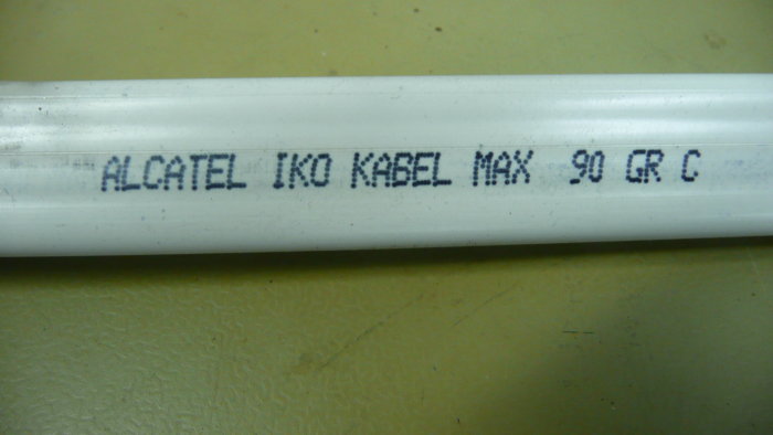 Närbild av en vit platt kabel märkt "ALCATEL IKO KABEL MAX 90 GR C" mot en gulaktig yta.