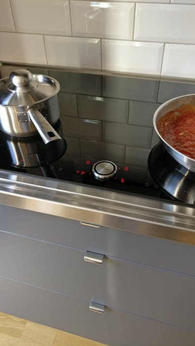 Induktionshäll med rostfri kastrull och tomatsås i en kastrull, mot en kaklad vägg och under köksskåp.