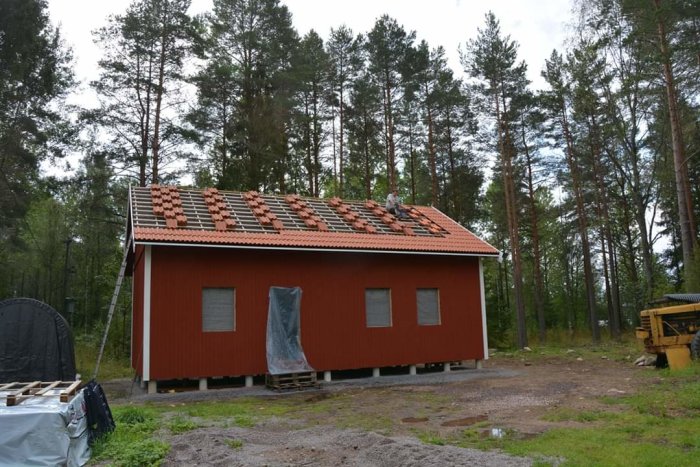 Rödmålat hus med tegeltak under konstruktion i skogsmiljö, person arbetar på taket.