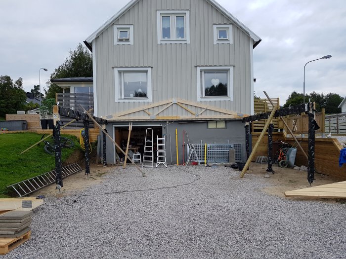 Nybyggd carport med öppen konstruktion framför ett hus, marken lutande, byggmaterial synligt.