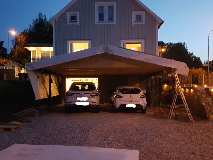 Nybyggd träcarport med plats för två bilar, täckt av vit presenning med bostadshus i bakgrunden.