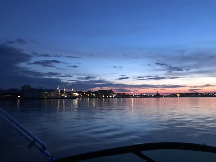 Solnedgång med rosa och blå himmel reflekterande över vattnet, sedd från en båt med stadssilhuett i bakgrunden.