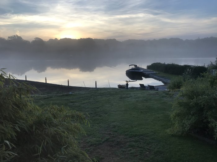 Morgondimma över ett lugnt vatten med båt vid brygga och soluppgång i bakgrunden.