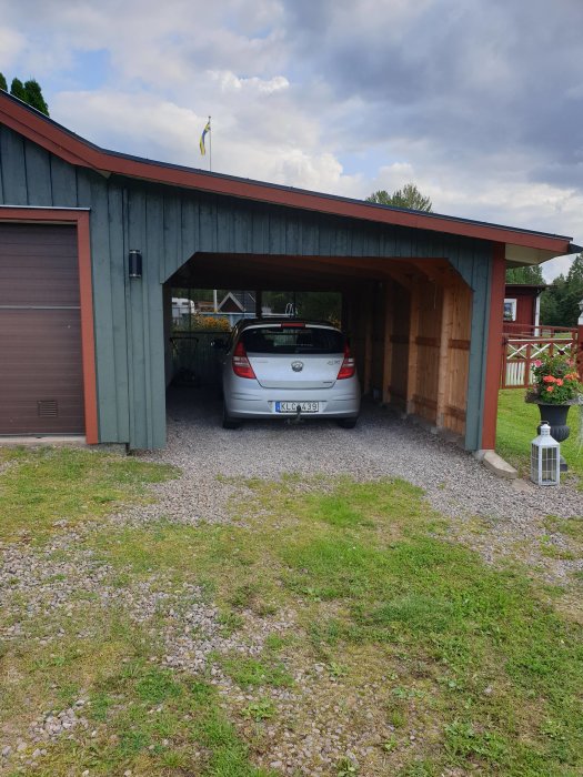 En bil parkerad under en carport med trasig takplåt som släpper igenom ljus, bredvid ett hus.