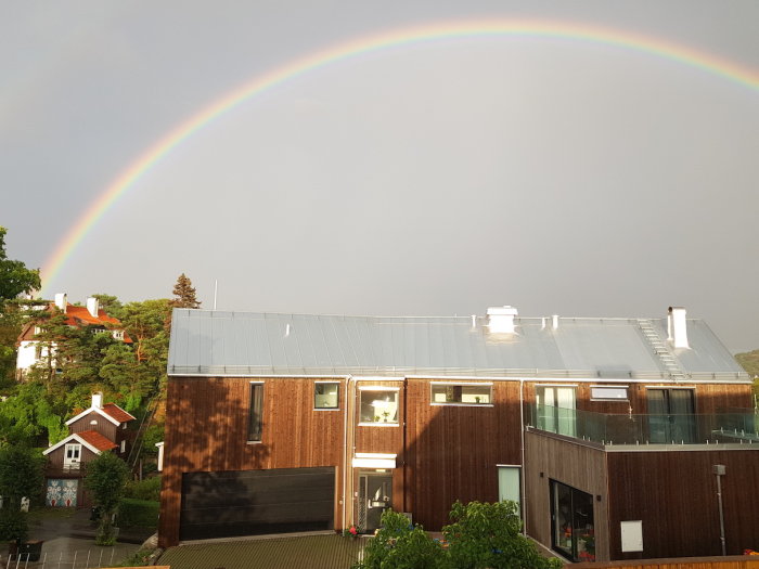 Regnbåge över ett vått hus med träpaneler under en regnskur.