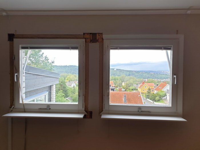 Två fönster med uppbrutna väggpartier ovanför, synliga reglar och isolering, utsikt över stad.