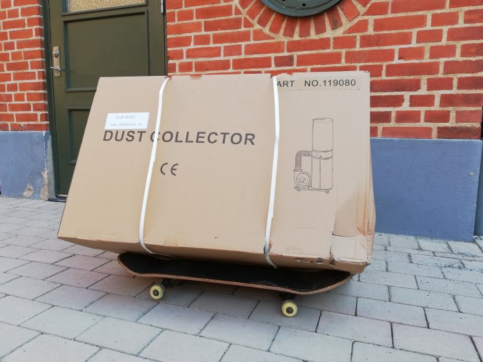 Stor kartong märkt "DUST COLLECTOR" på en skateboard framför en tegelvägg och grön dörr.